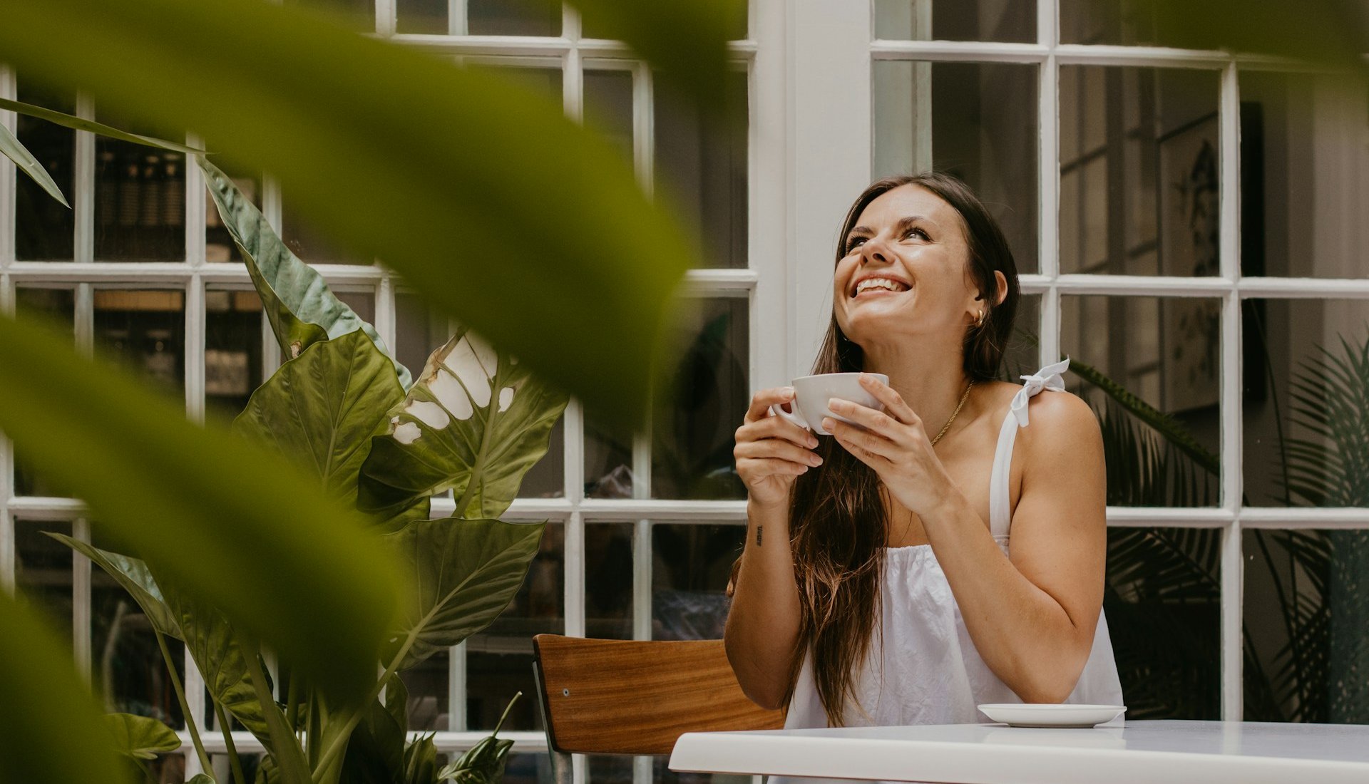 Mulher de cabelos longos sorri olhando para cima, segurando uma xícara de café com as duas mãos, sentada à mesa de um café com plantas ao redor