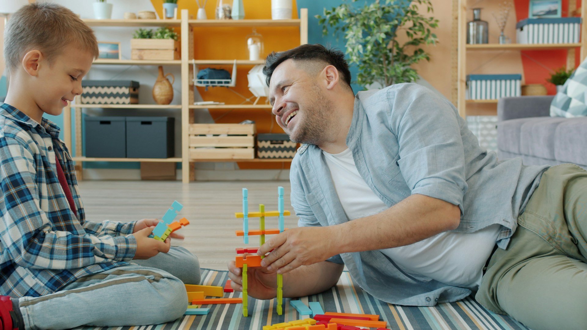 Pai e filho sorriem sentados no tapete da sala, montando uma torre com peças coloridas de encaixe