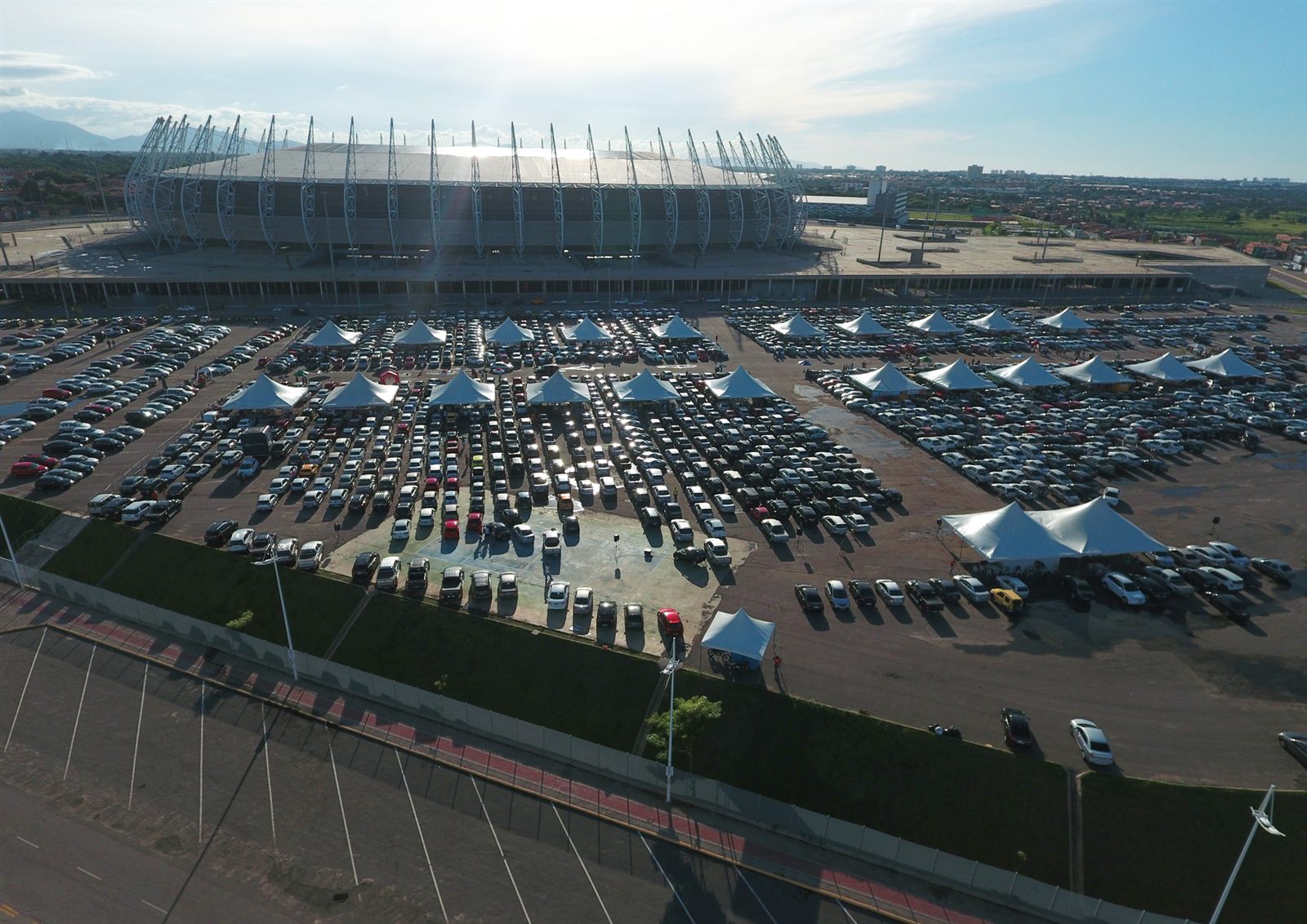 Vista aérea de um feirão de veículos, com centenas de carros enfileirados e tendas brancas ao lado de um estádio