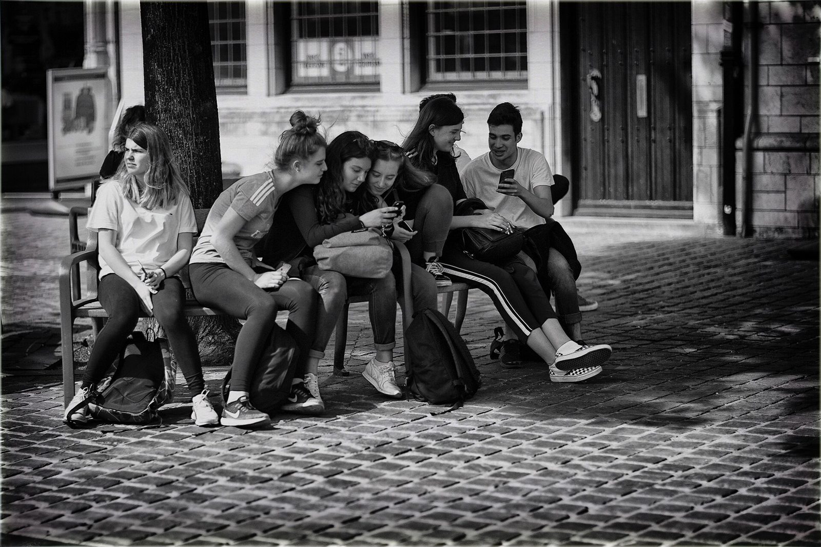 Fotografia em preto e branco de adolescentes em um banco de praça, inclinados sobre as telas de dois celulares
