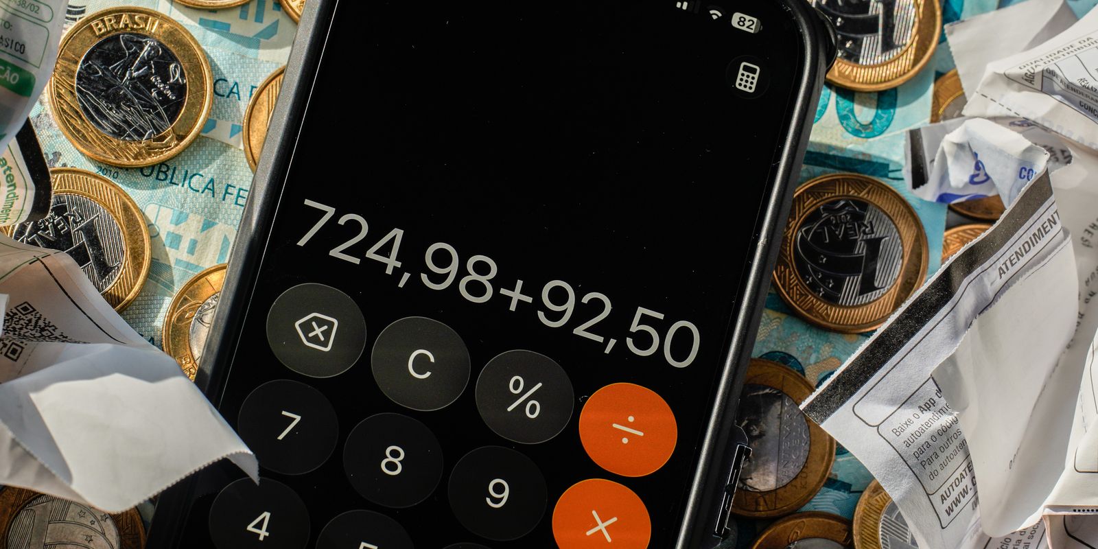 Celular com a calculadora aberta sobre moedas e notas de real e cupons de pagamento amassados