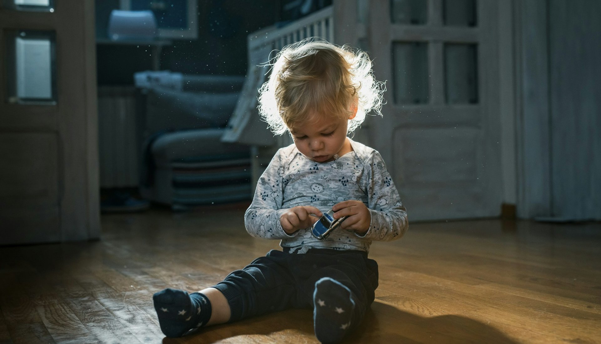 Criança pequena de cabelo claro, sentada no chão de madeira de uma casa à meia-luz, brinca concentrada com um carrinho de brinquedo