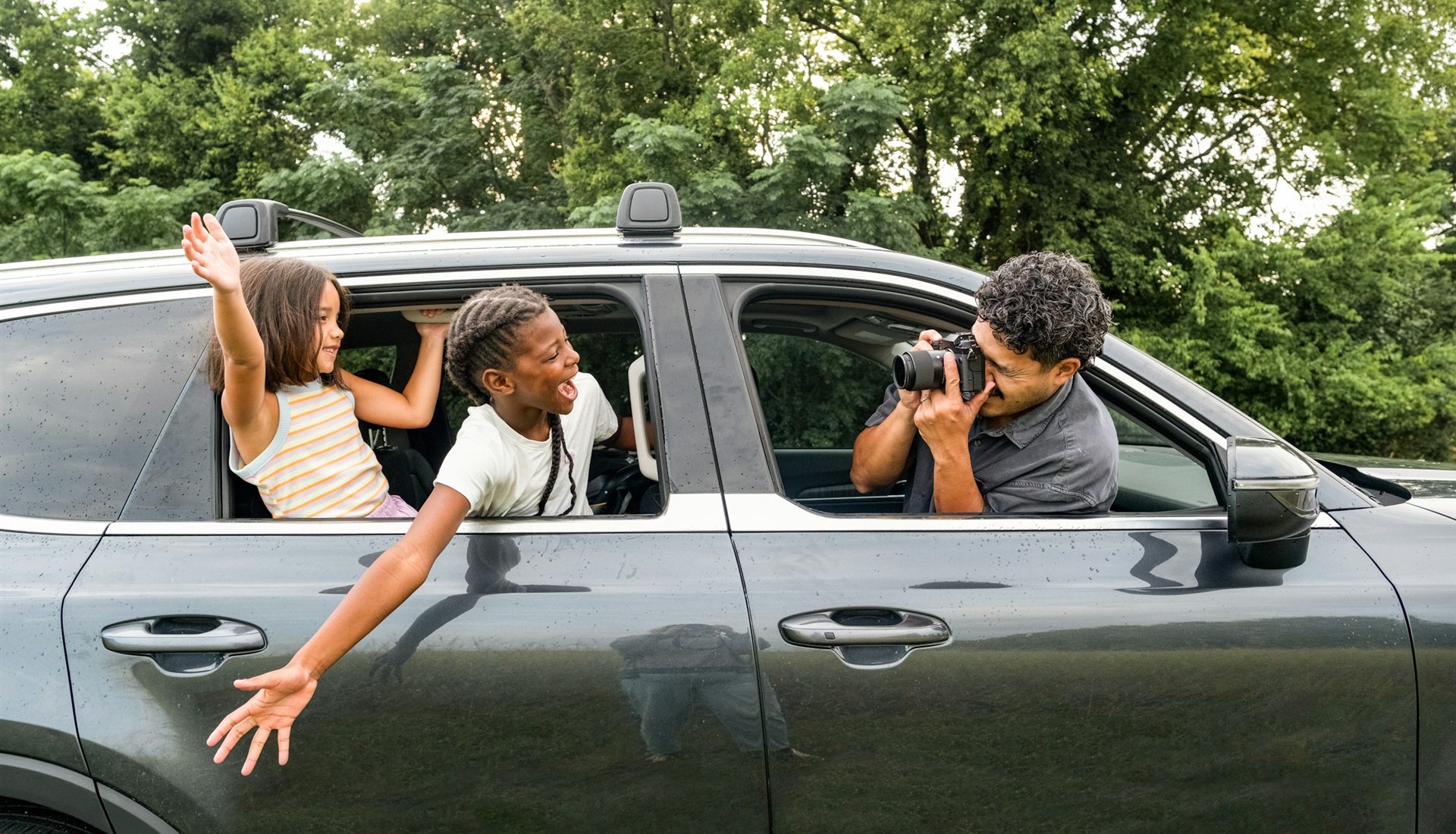 Duas meninas sorriem e acenam com os braços para fora da janela traseira de um carro escuro, enquanto um homem, no banco da frente, as fotografa pela janela