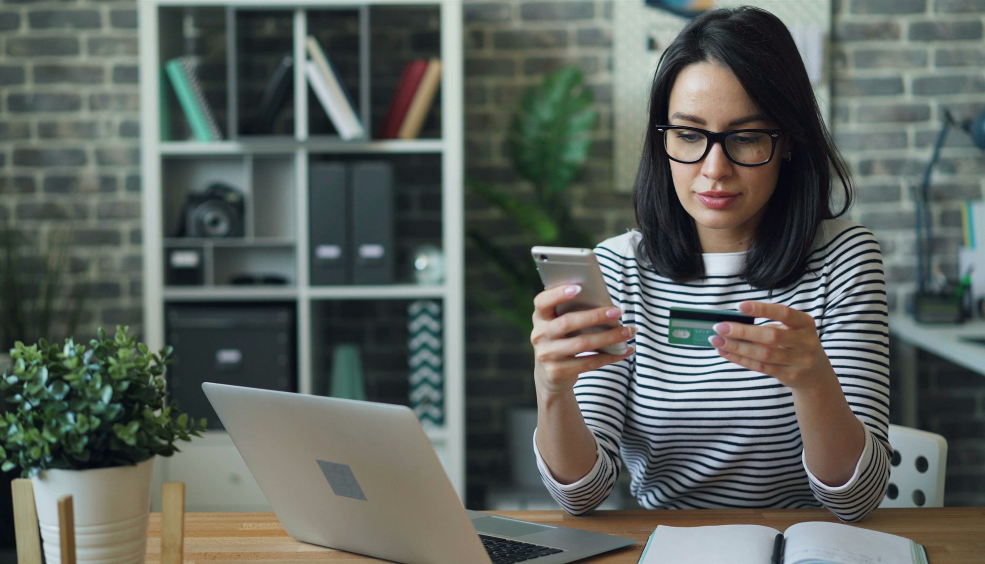 Mulher de óculos confere o celular com um cartão na mão, diante de um notebook