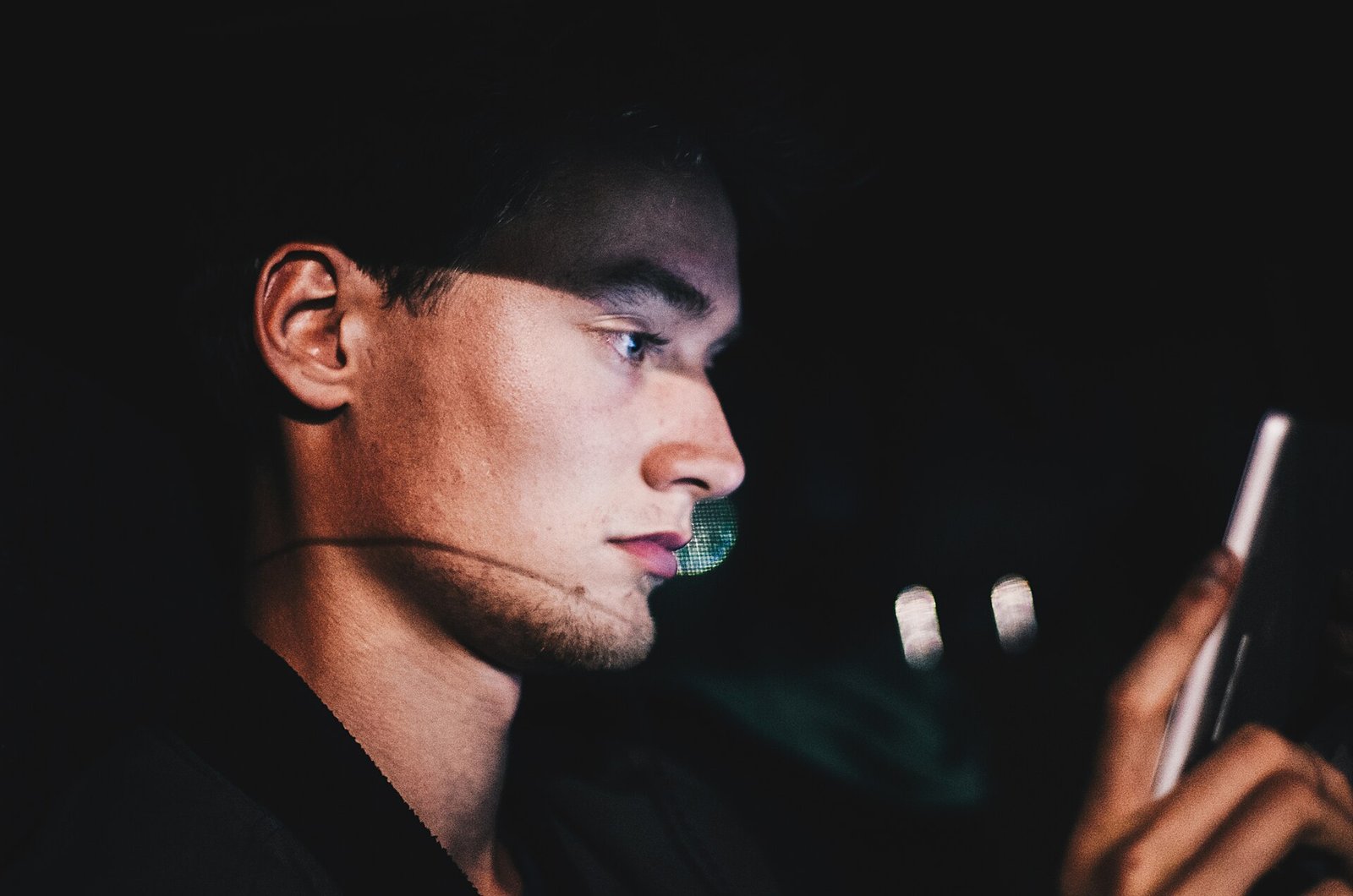 Rosto de um homem jovem, de perfil, iluminado no escuro pela tela do celular