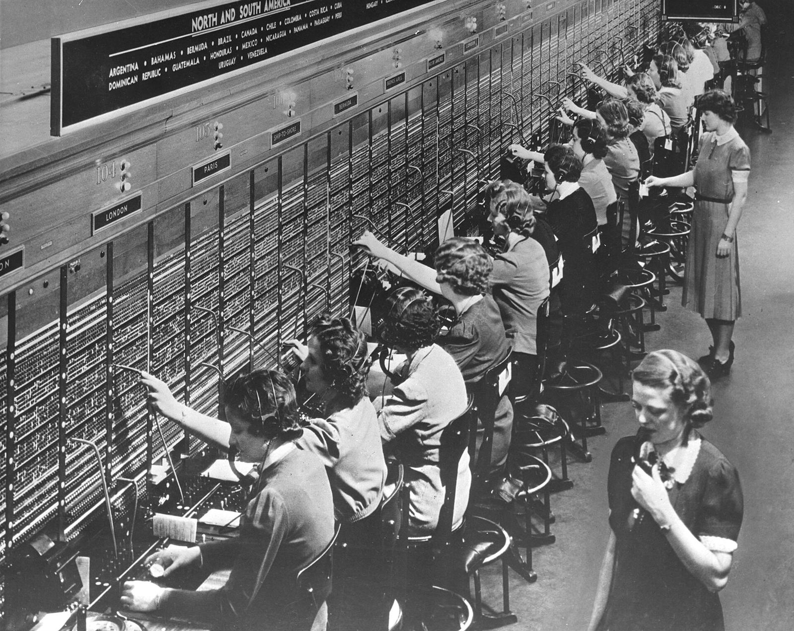 Fotografia em preto e branco de uma fileira de telefonistas diante de um painel de conexões, na década de 1940