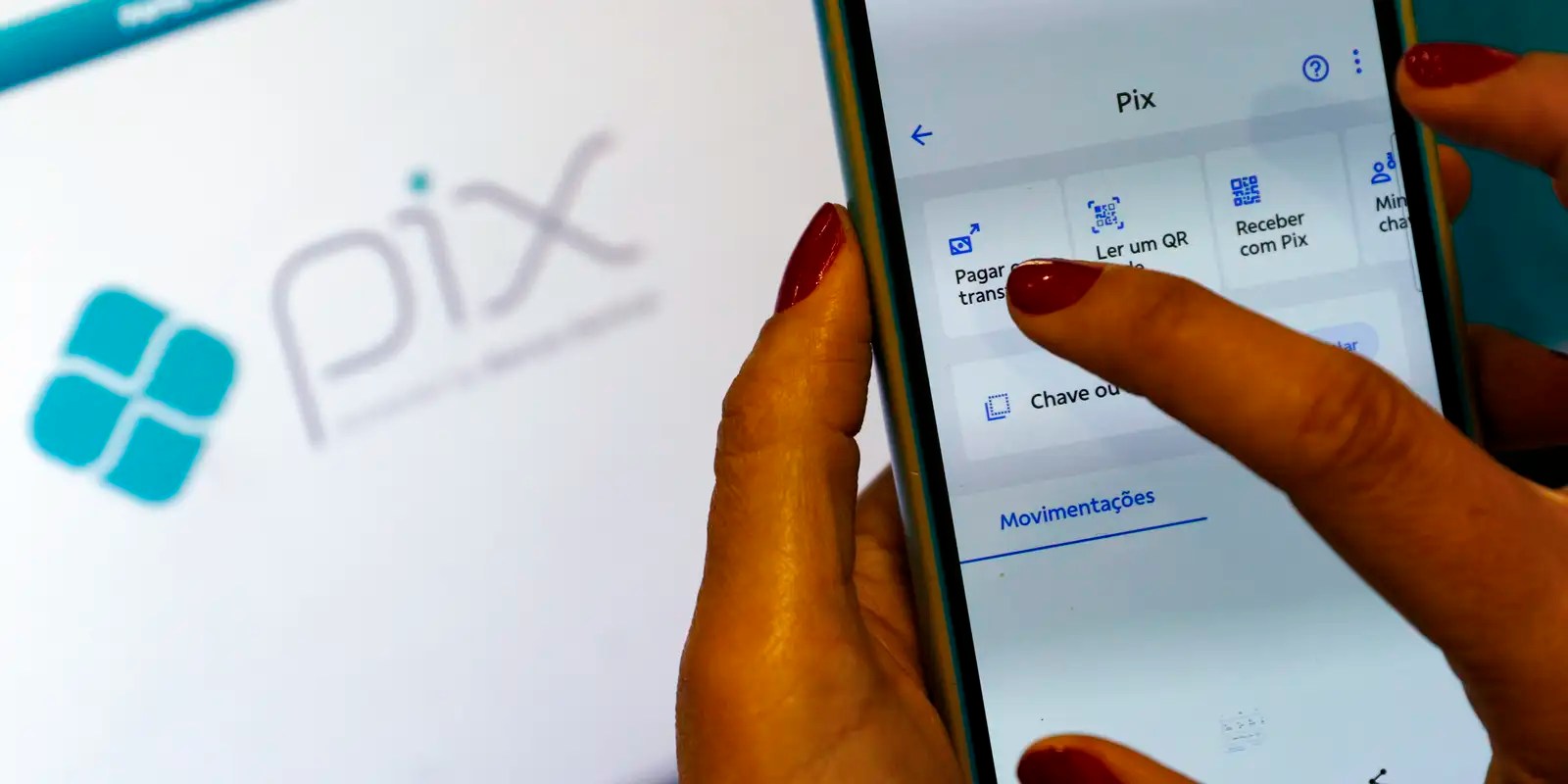 Mão com unhas vermelhas toca a tela de um celular aberto na área Pix do aplicativo de um banco; ao fundo, desfocada, a marca do Pix