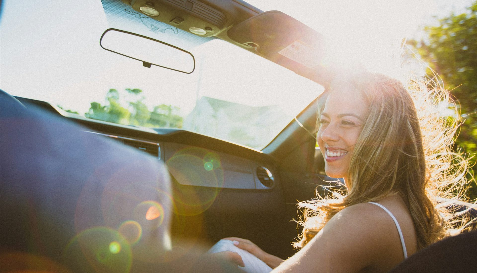 Jovem de cabelos longos sorri no banco de um carro conversível, com o cabelo ao vento e o sol entrando pelo para-brisa