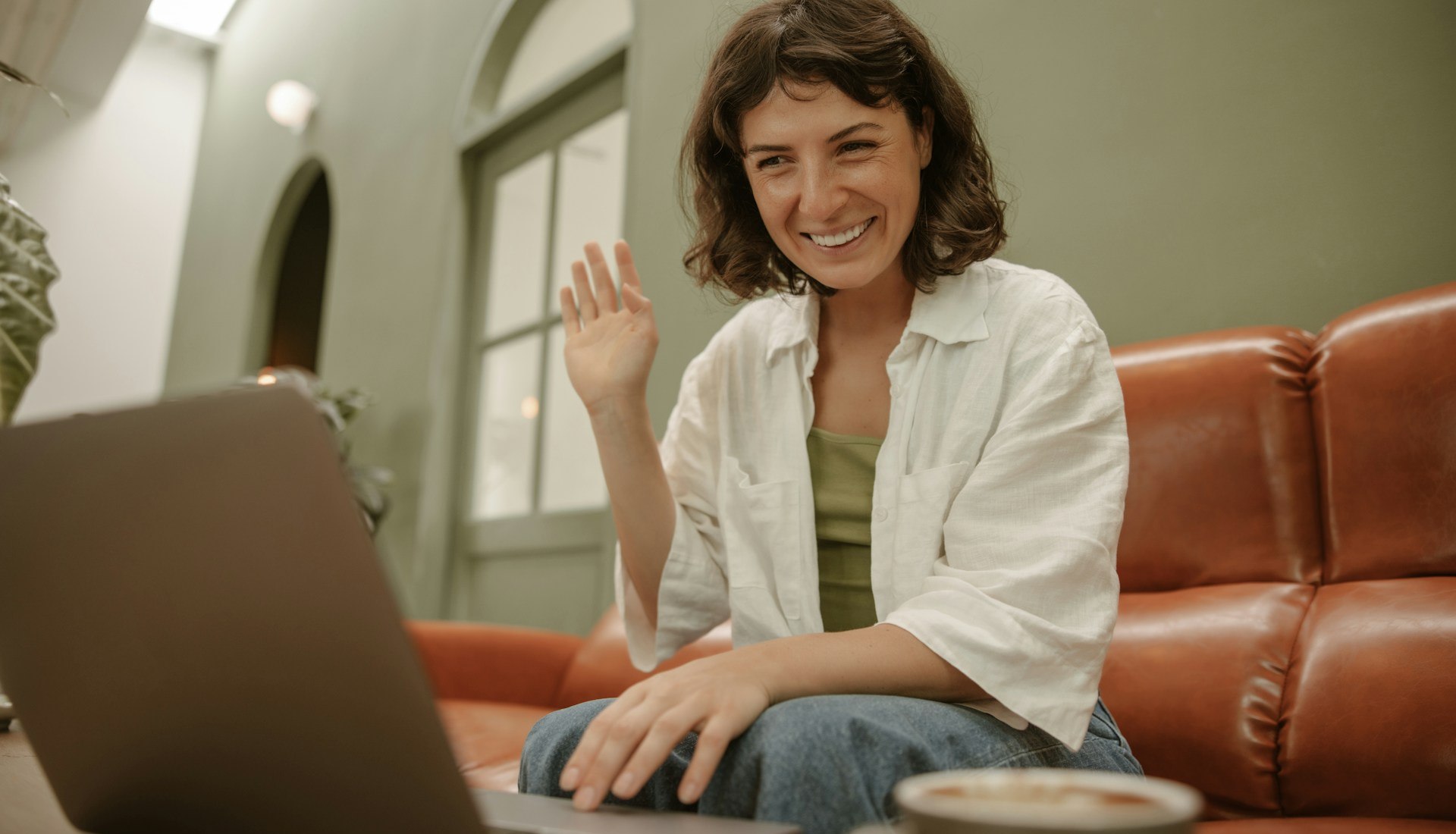 Mulher de cabelo curto e camisa branca, sentada em um sofá de couro, sorri e acena para a tela de um notebook aberto à sua frente