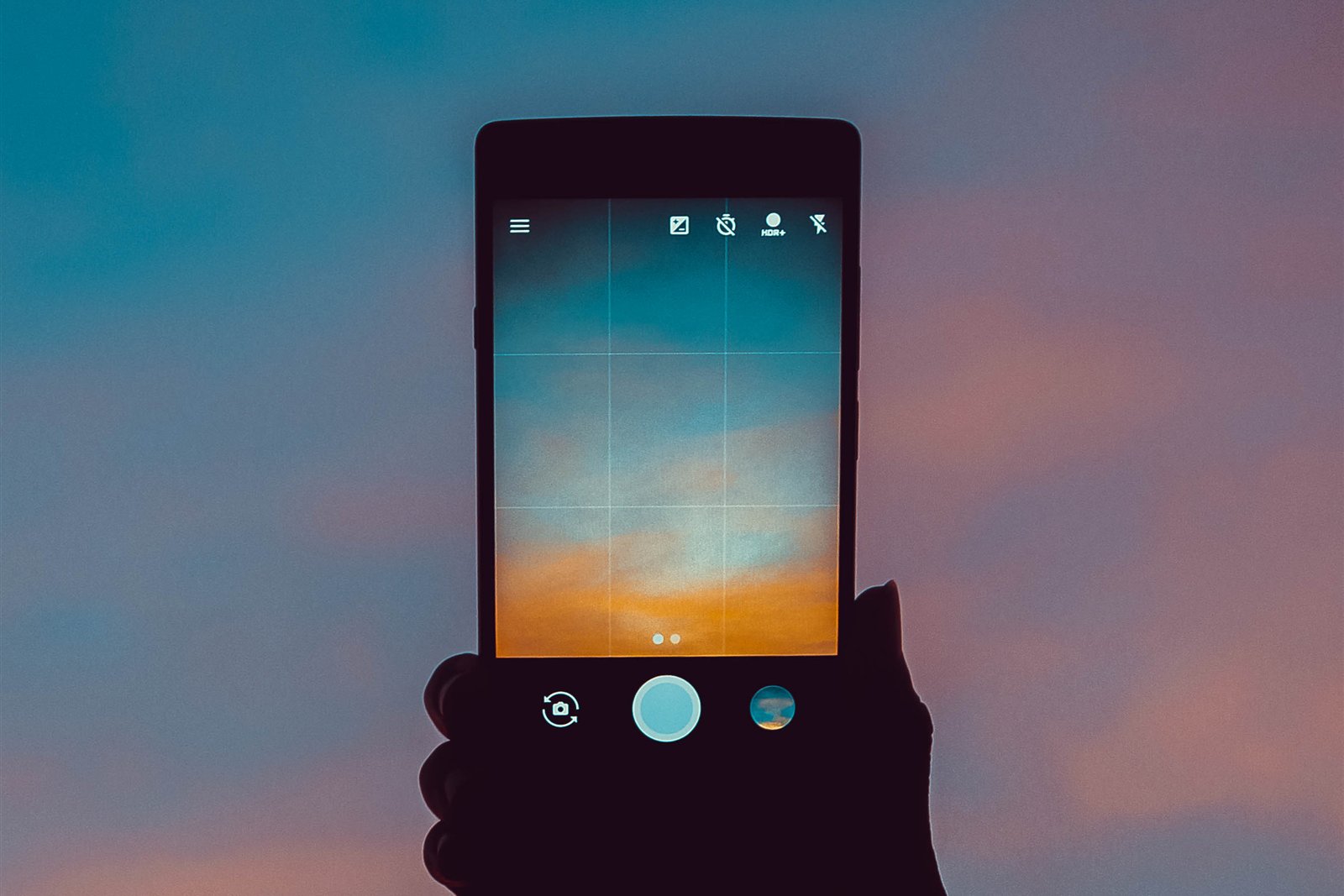 Mão em silhueta segura um celular contra o céu do fim da tarde, com a câmera aberta na tela